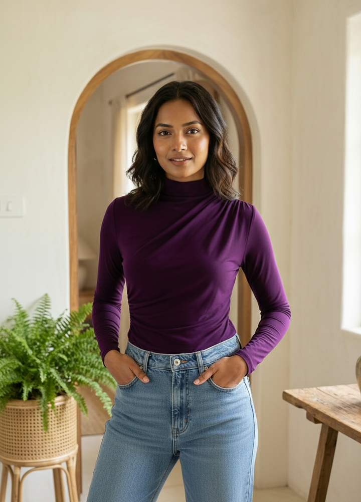 Woman wearing a purple turtleneck and blue jeans standing in a room with a mirror and plant.